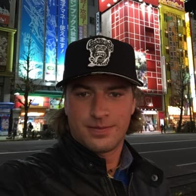 Man in a black cap and leather jacket poses before vibrant neon city signs.